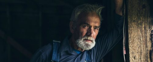 An older man with gray hair and beard, wearing a blue shirt and overalls, leans against a wooden post in a dimly lit setting, reflecting the quiet wisdom found in many Reca portraits.