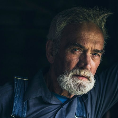 An older man with gray hair and beard, wearing a blue shirt and overalls, leans against a wooden post in a dimly lit setting, reflecting the quiet wisdom found in many Reca portraits.