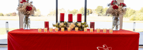 A table covered with a red cloth holds multiple red and white candles and two floral arrangements in jars, set in front of a large window with a view of trees and water.