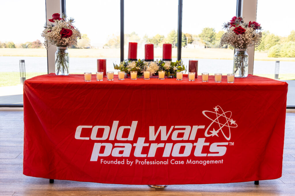 A red tablecloth with "Cold War Patriots" and "Founded by Professional Case Management" on it, decorated with candles, flowers, and red vases, honors the National Day of Remembrance in front of large windows.