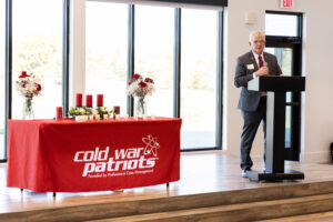 Gary Wilson speaks at a podium next to a table with red candles and flowers, covered by a red "Cold War Patriots" tablecloth in a bright room.