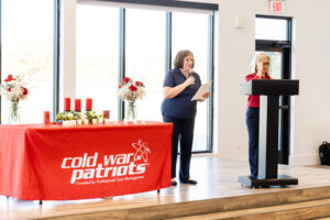 Nikki Clark and Judy Terry stand at the front of a room, one speaking at a podium and the other holding a microphone beside a table with a red "Cold War Patriots" tablecloth and candles.
