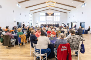 A large group of people sit at round tables in a bright banquet hall, facing a stage where a choir is performing.
