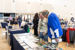 Two older men look at a table covered with photos and documents at an indoor event with other people seated and walking in the background.