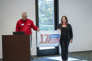 Two people stand indoors holding a "17 Years, Cold War Patriots National Day of Remembrance" banner; one person is at a podium, and a door labeled "Oak Ridge Conference Center" is in the background.