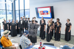 A choir of young adults in formal black attire sings under a conductor in front of an audience, with a large TV screen displaying the event logo in the background.