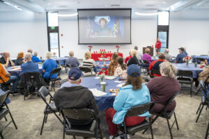 A group of people sits at round tables in a conference room, watching a video presentation from the U.S. DOL Ombudsman.