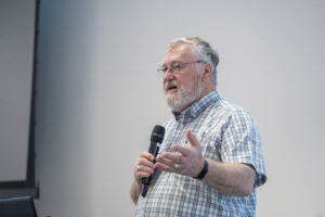 Ray Smith speaks into a microphone while gesturing with his hand, standing indoors against a plain light-colored background.