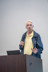 Larry Washam stands behind a podium, holding a microphone and speaking, with a laptop in front of him.