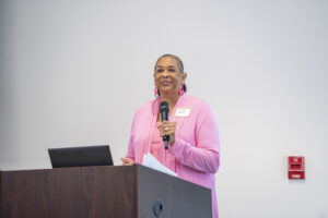 Connie Dunn stands at a podium, holding a microphone and speaking in front of a plain white wall.