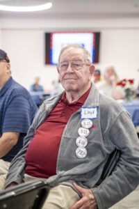 Lloyd sits indoors, with several event buttons pinned to his jacket. Other people and a screen are visible in the background.